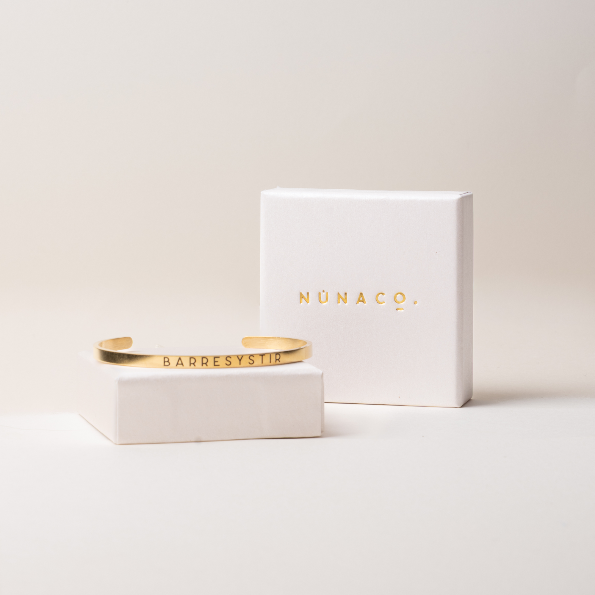 A gold cuff bracelet engraved with 'BARRESYS19' displayed on a small white pedestal next to its white presentation box with gold foil 'NUNACO' branding, set against a warm off-white background.