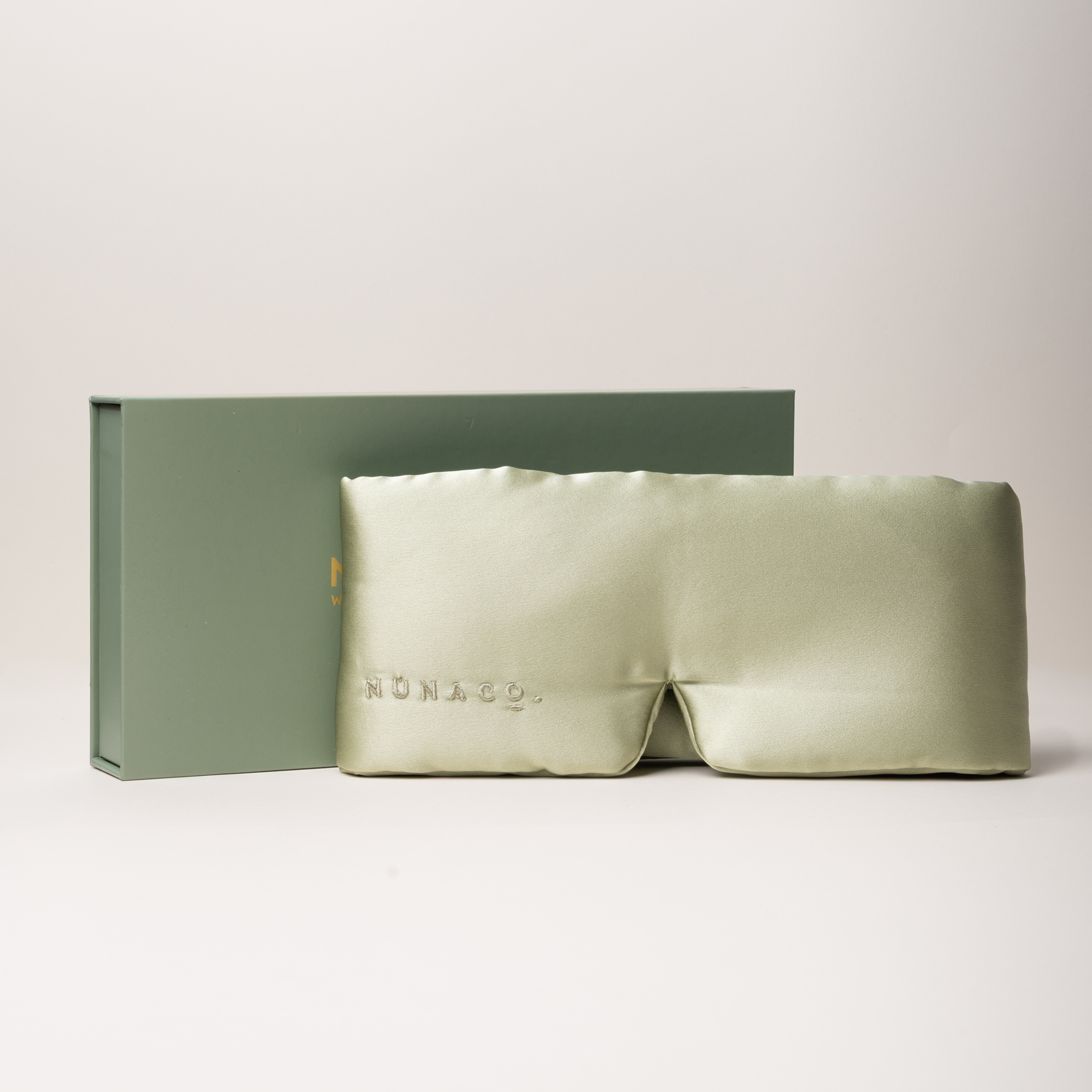 A pale sage green satin sleep mask with 'MUNARO' debossed on the front, displayed in front of its matching sage green presentation box on a clean, light beige studio background.