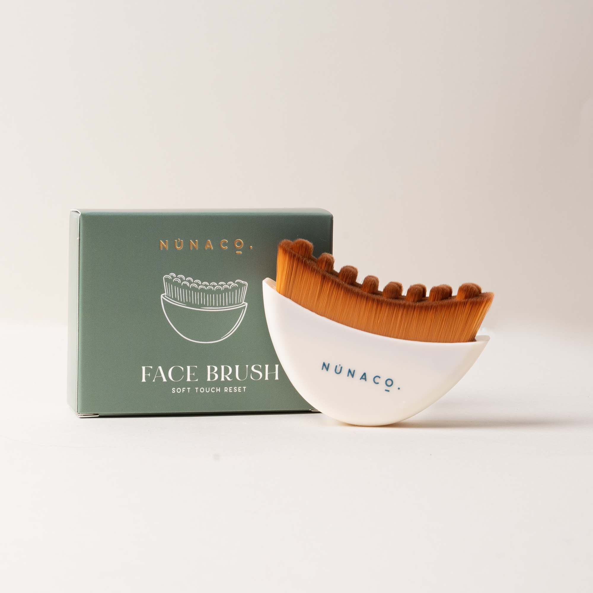 Nunaco face brush set with a white crescent-shaped holder, brown bristles, and a sage green product box on a light beige background.