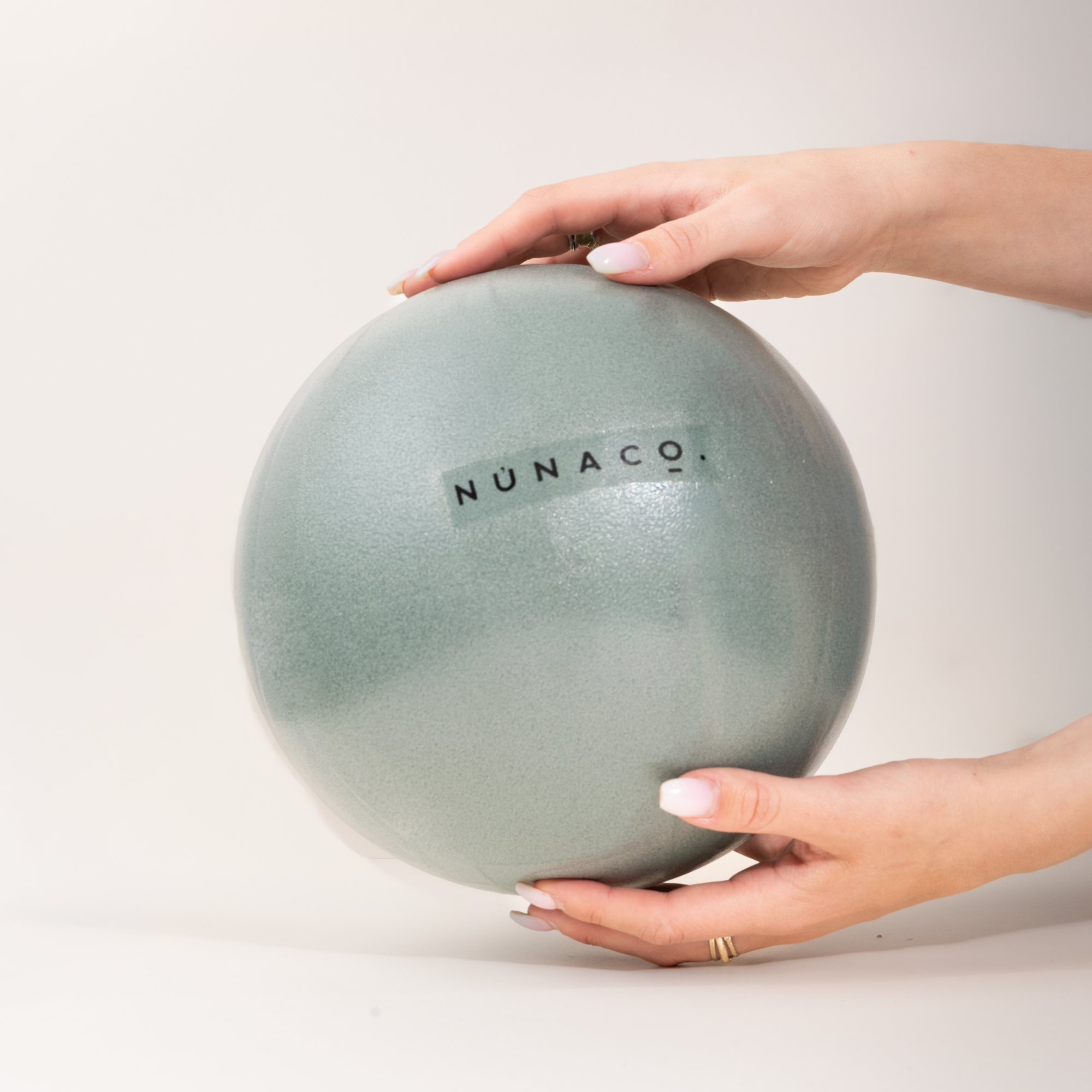 A person's hands holding a matte sage green ceramic sphere with the brand name 'NUNACO' printed on it, set against a clean, light off-white background.