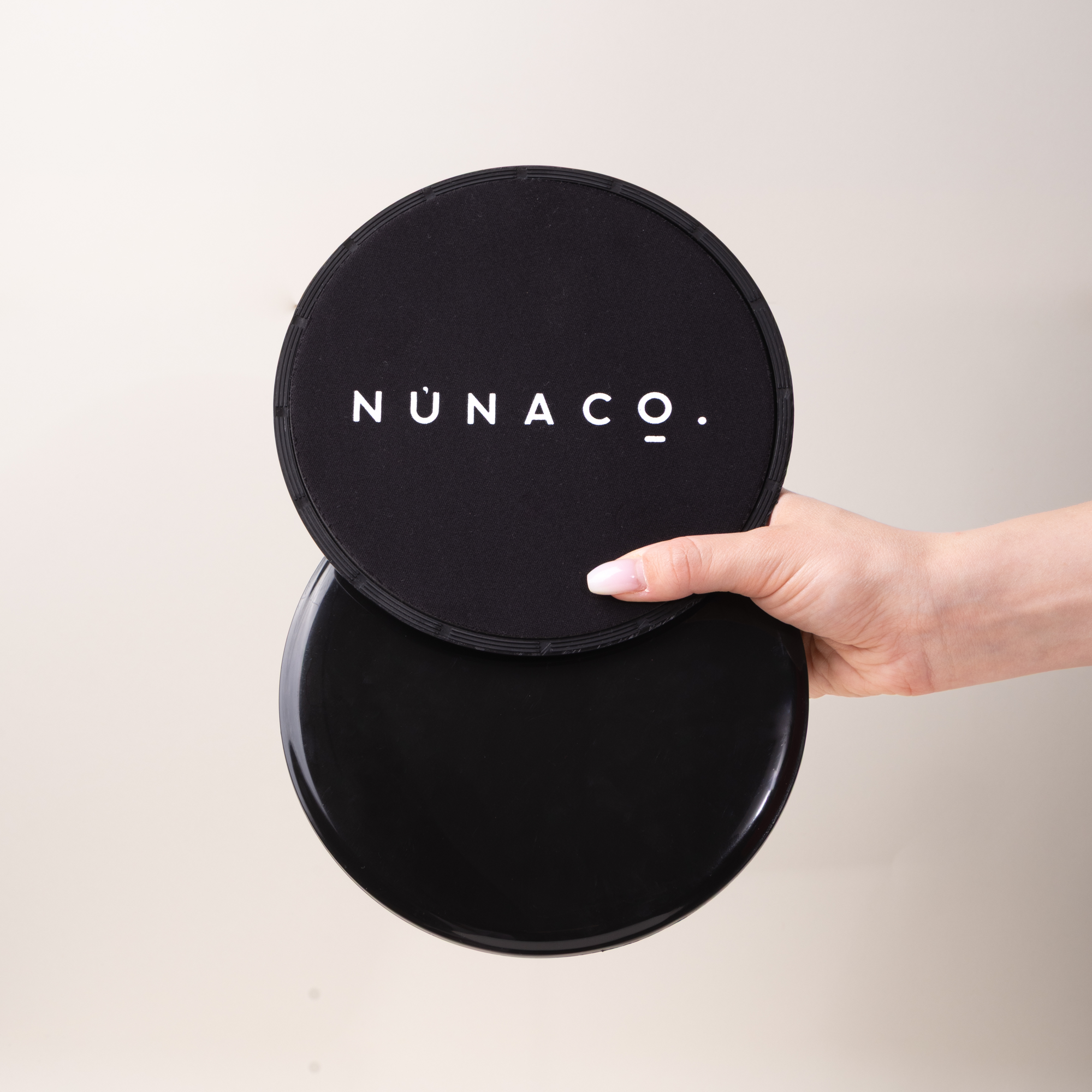 A hand holding a round black cushion with the brand name 'NÜNACO.' printed in white on the top cushion, set against a plain light beige background.