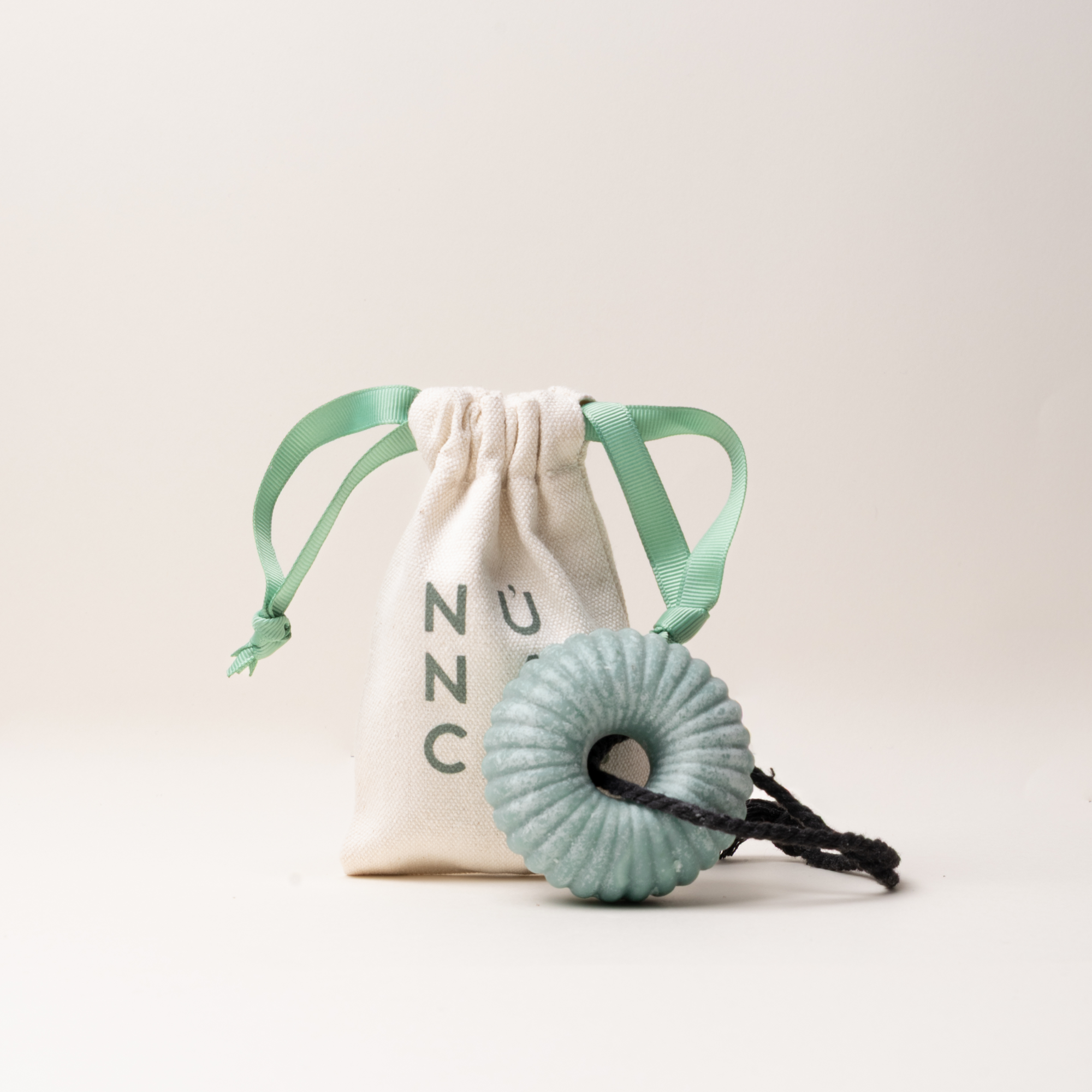 A mint green ribbed donut-shaped massage roller with a black cord, displayed in front of a cream-colored drawstring pouch with green ribbon ties and 'UNC' printed on the front, set against a solid off-white background.