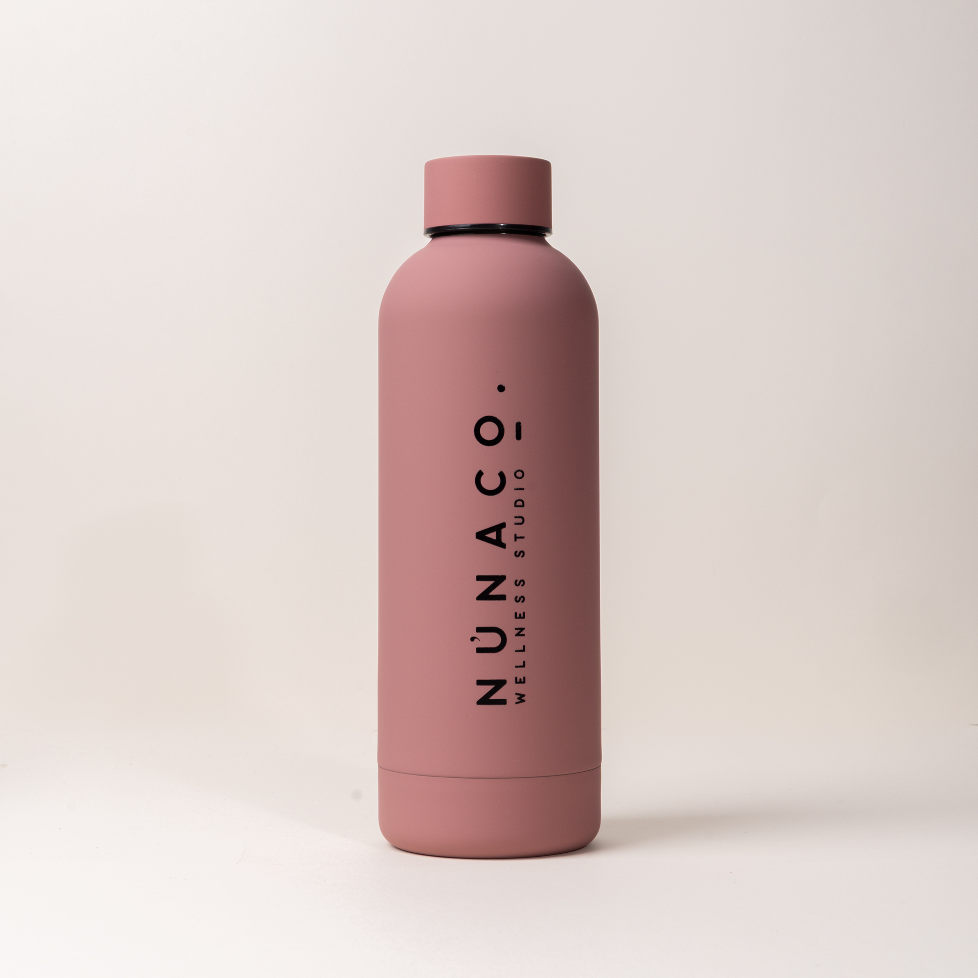 A matte dusty pink insulated water bottle with a matching pink lid, featuring the vertical black text 'NUNACO. WELLNESS STUDIO' on the front, set against a clean off-white background.