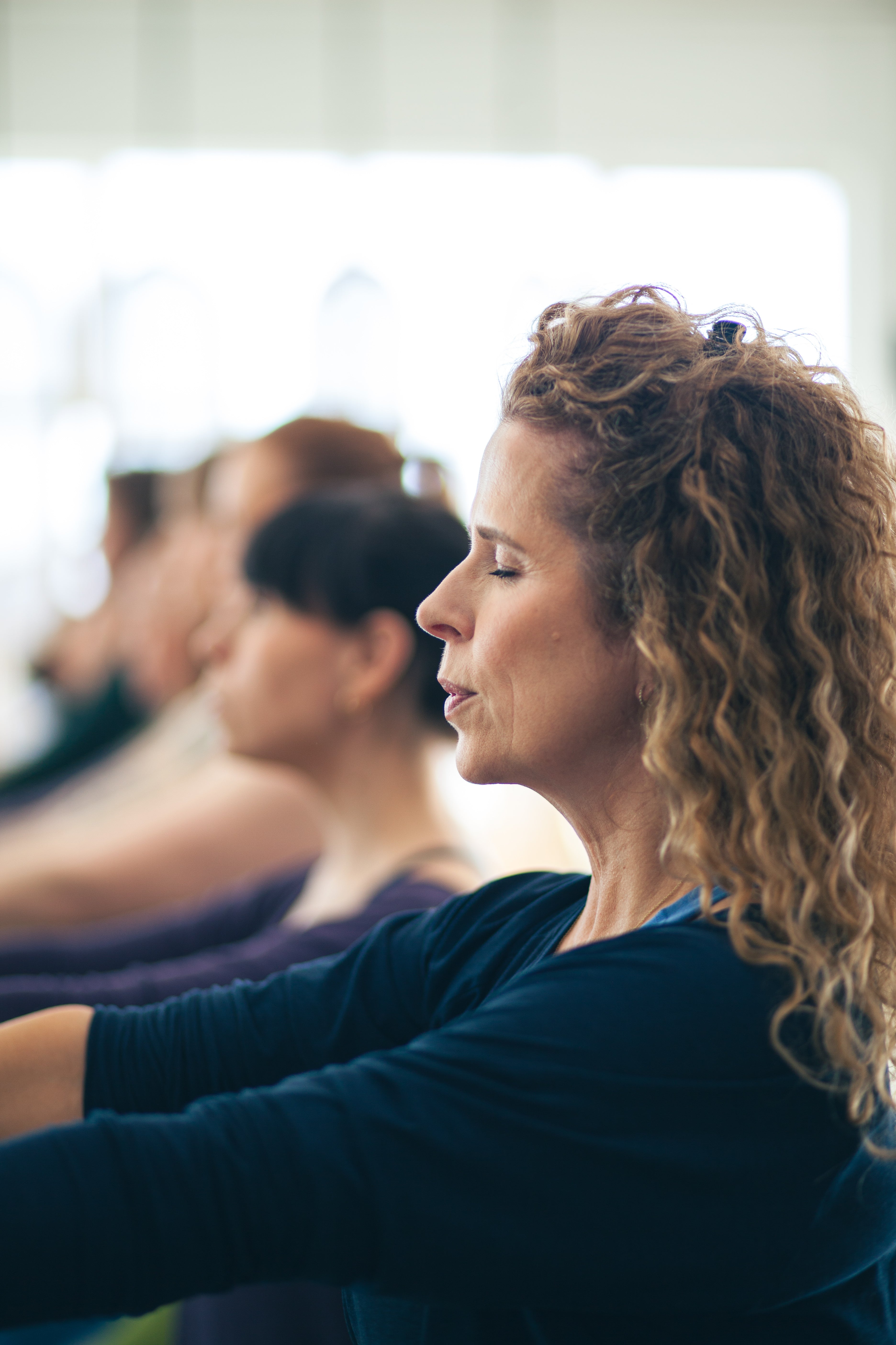 A woman with curly light brown hair in profile, wearing a dark navy long-sleeve top, sitting in a yoga or meditation class with other participants blurred in the background.