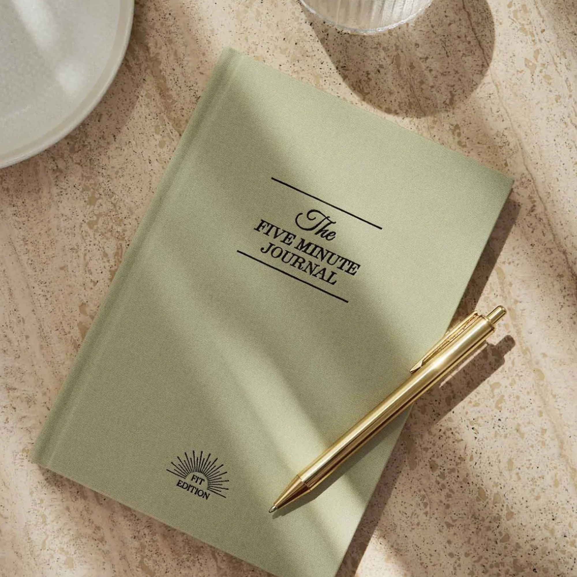 A sage green hardcover journal titled 'The Five Minute Journal' lies on a beige stone surface, with a gold pen resting on top and partial white dishes visible at the edges.