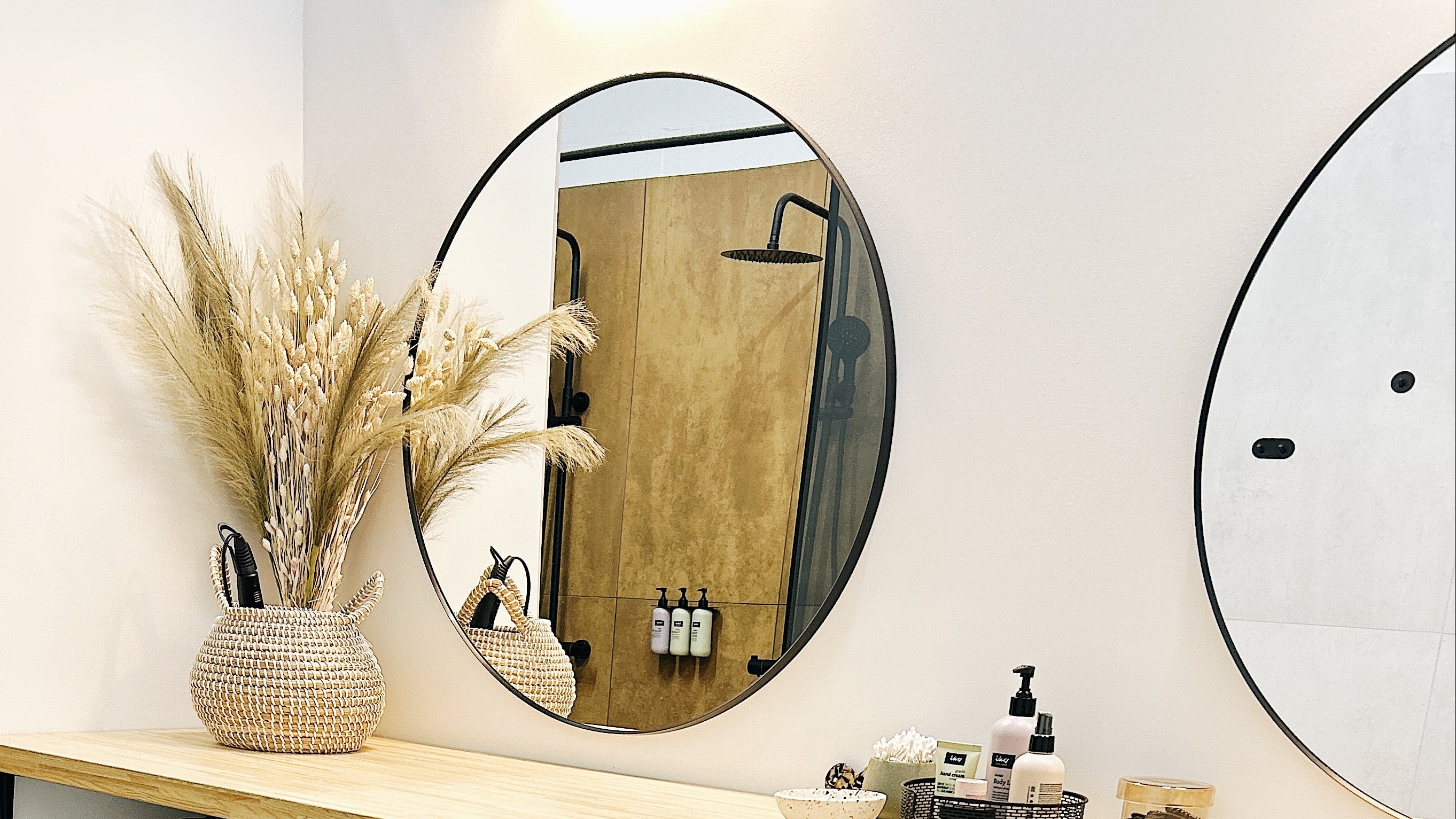 A spa or bathroom vanity setup with a light wood shelf holding toiletries and dried grass in a woven vase, two round wall mirrors, and multiple wicker baskets filled with folded green towels or linens and white rolled towels.
