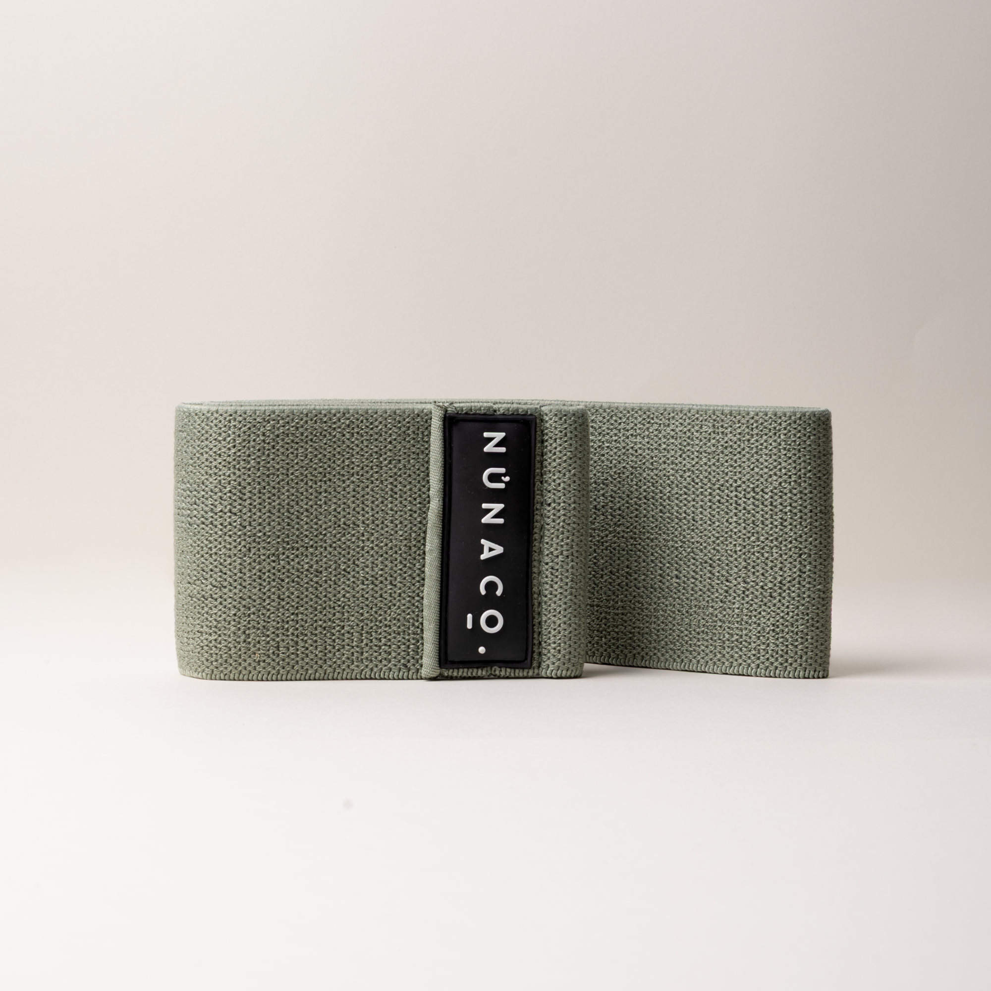 An olive green textured fabric strap with a black vertical label reading 'UNACO' in white, photographed on a clean light gray studio background.