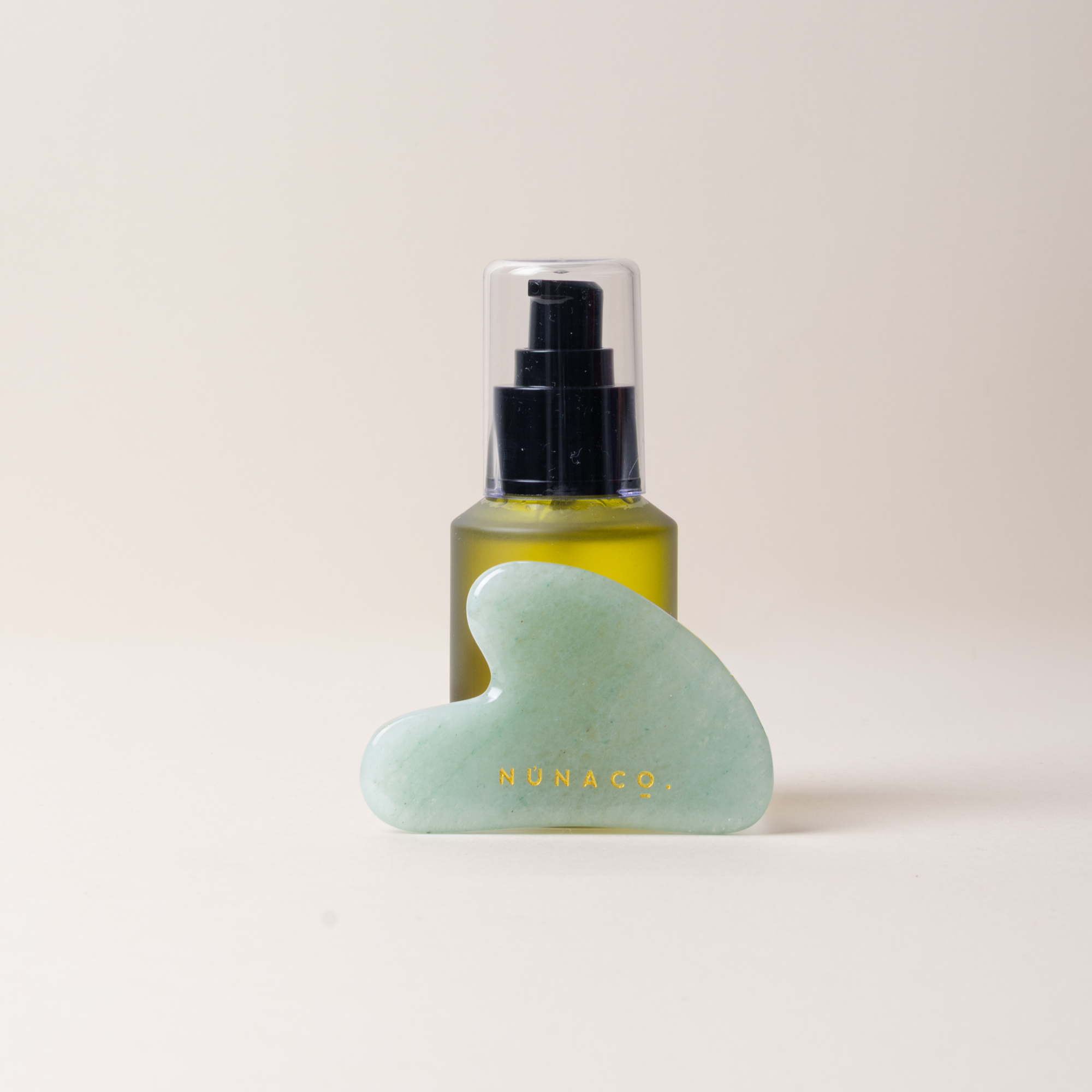 A light green jade Gua sha massage tool with gold lettering, positioned in front of a small amber spray bottle with a black pump and clear cap on a warm off-white background.