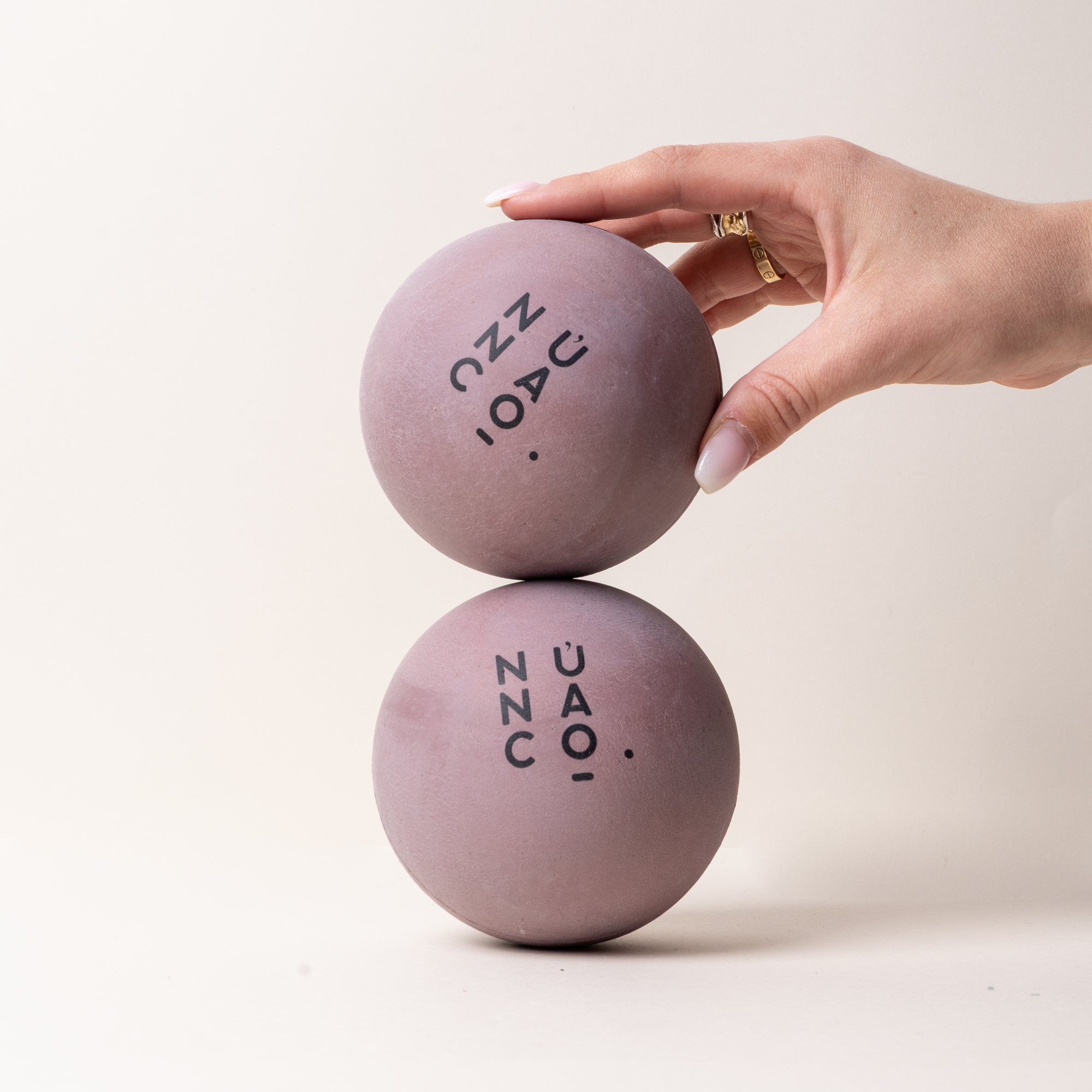 Two dusty purple foam balls with black text printed on them, stacked with a hand holding the top one against a plain off-white background.