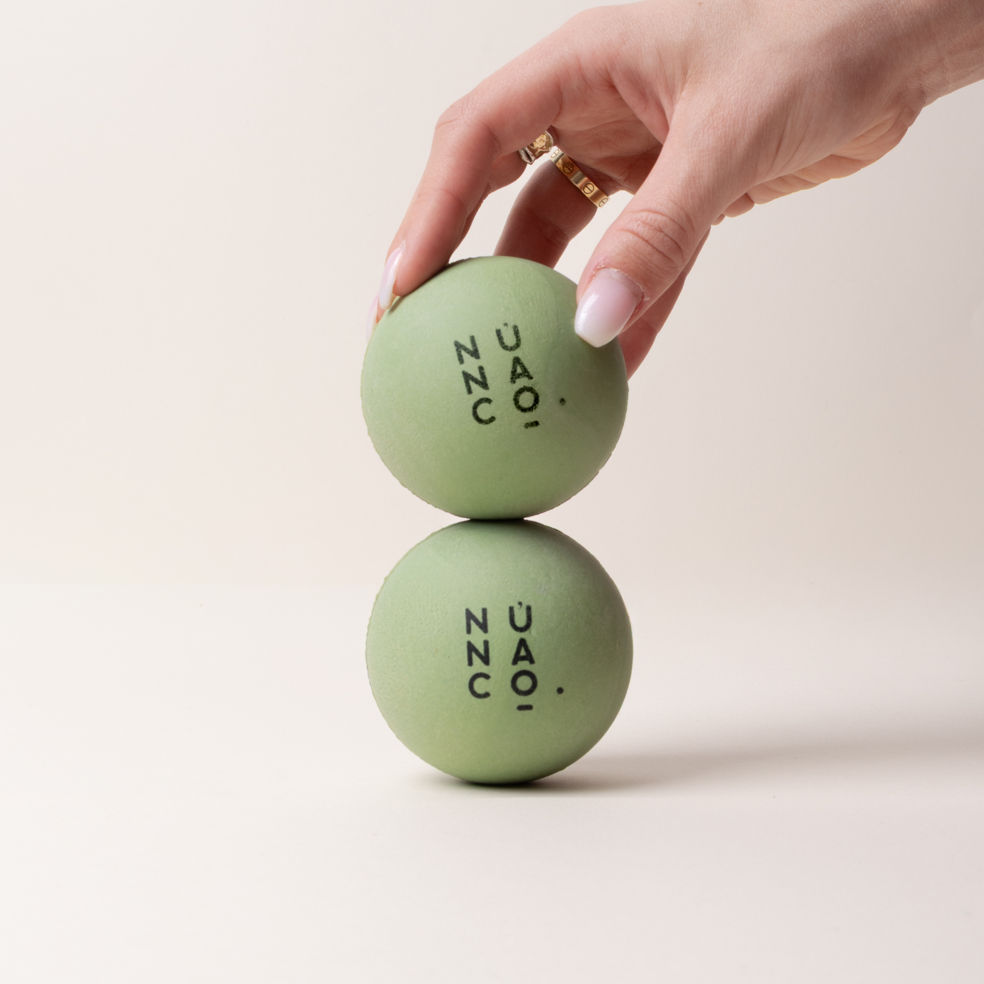 A hand with a gold ring holds a light sage green stress ball above another matching ball on a clean, light beige background. 