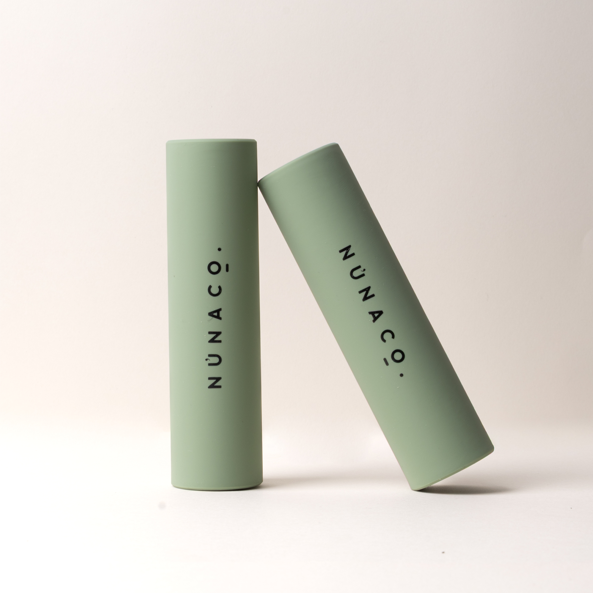 Two sage green cylindrical cosmetic tubes standing upright on a light beige studio background, with the brand name NUNACO printed vertically in black on each tube.