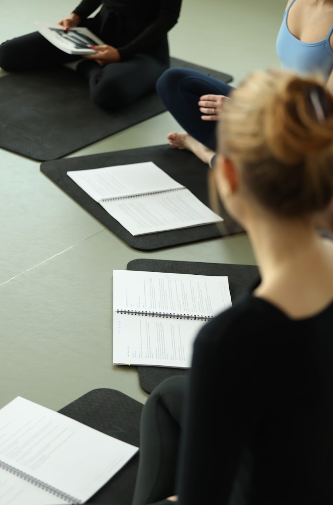 Several people sitting cross-legged on black exercise mats in a studio, each with an open notebook in front of them. The foreground shows the back of a woman with her hair in a bun wearing a black top, and another person in a light blue top is visible in the upper right.