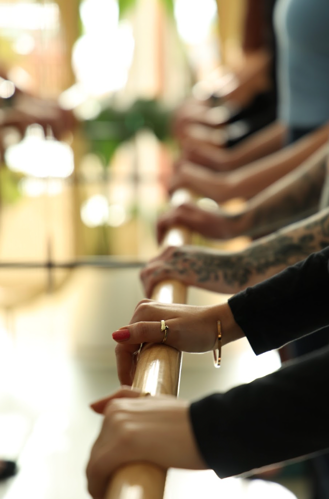 Close-up of multiple people's hands gripping a wooden ballet barre in a studio, with shallow depth of field and warm, bright background lighting.