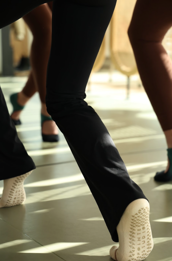 Close-up of a person's legs wearing black pants and white textured non-slip socks or slip-on shoes, standing on a glossy floor with sunlight and shadows.