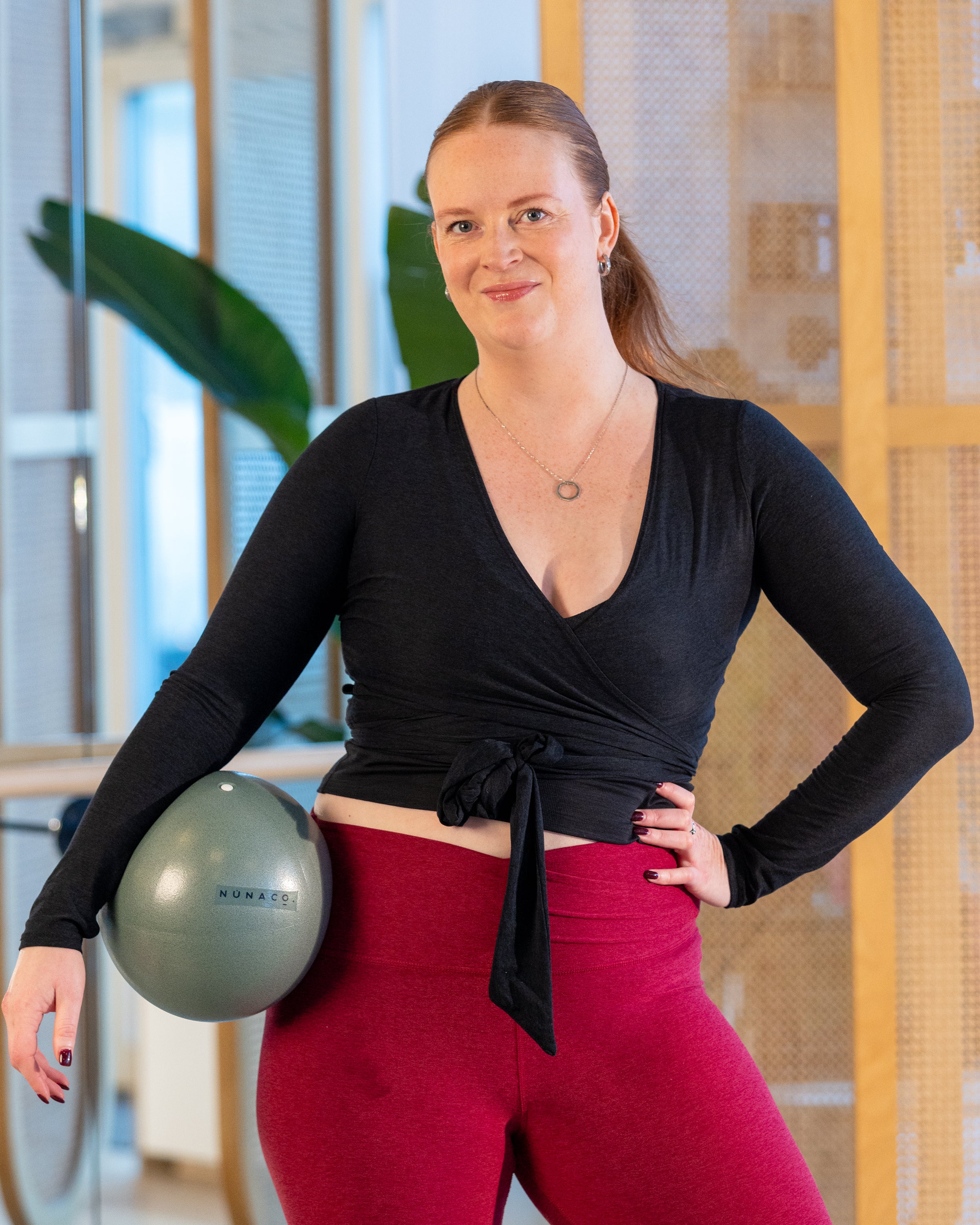 Woman in black long-sleeve top and red leggings holding a silver exercise ball indoors.