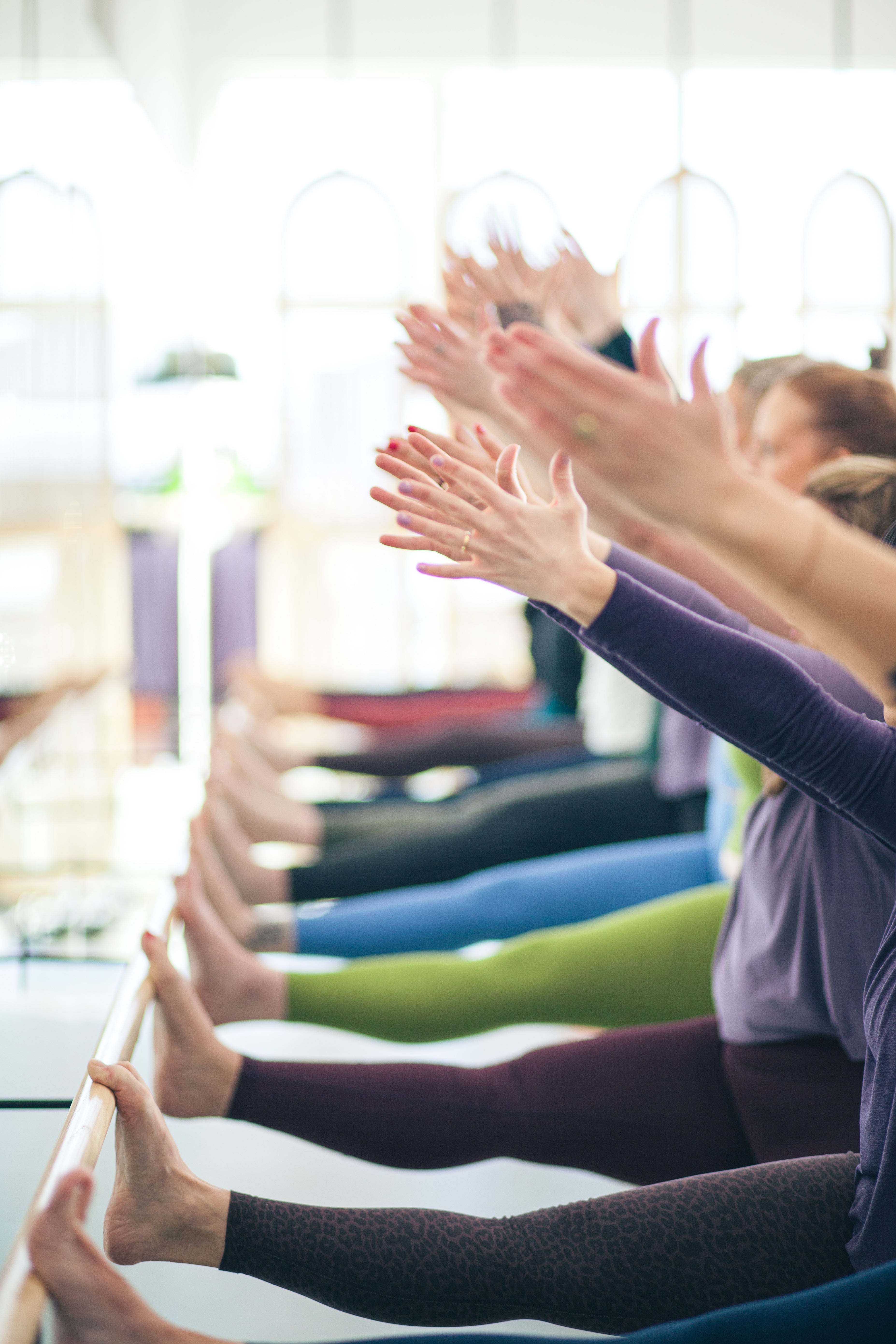 A row of people in a yoga studio performing a seated pose with arms raised, wearing colorful leggings in purple, green, blue, and black.