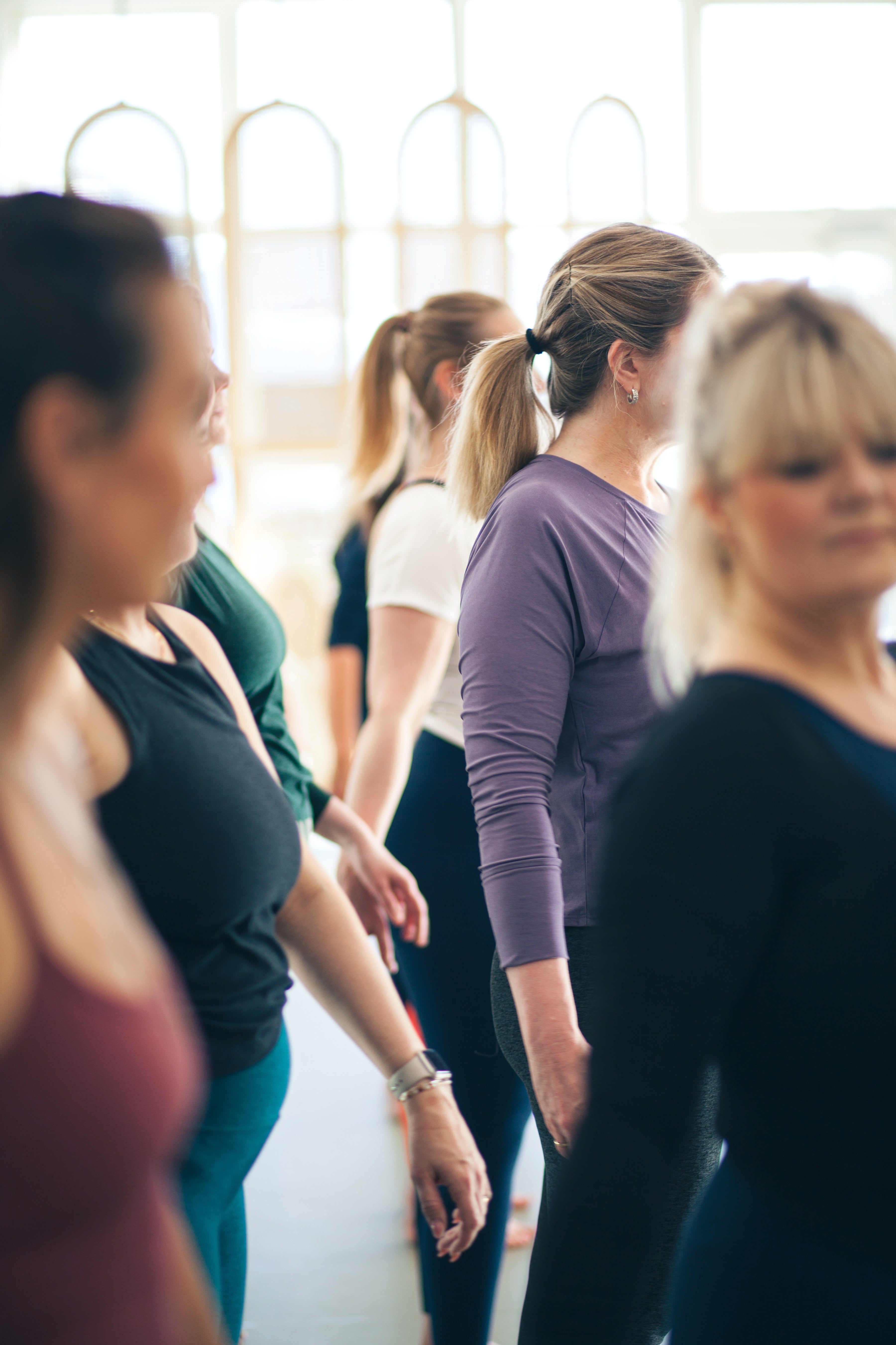 A group of women in a bright studio or gym, standing in a line. The central figure wears a purple long-sleeve athletic top and black leggings, with other participants in dark tops and leggings visible around her.