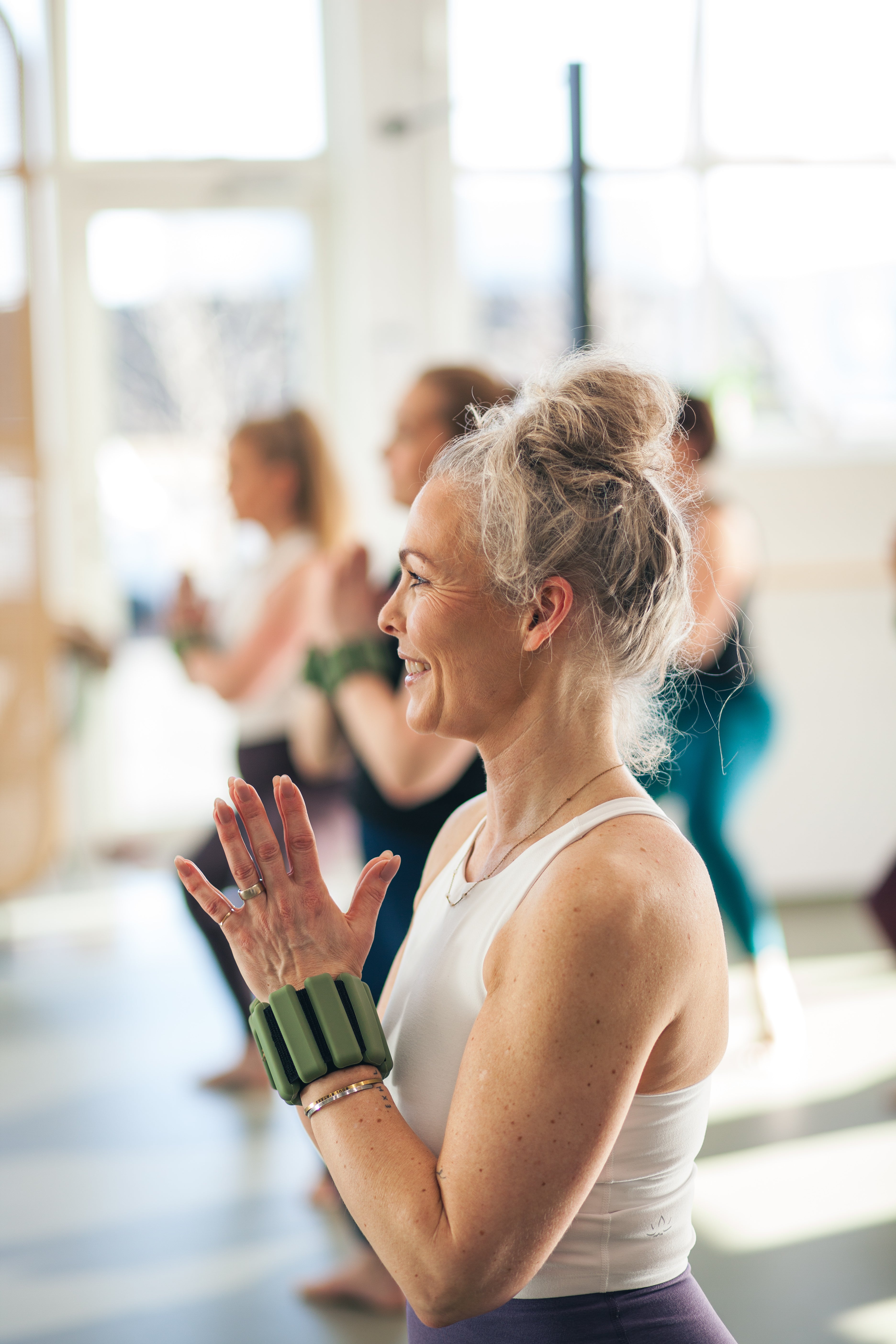 A woman in a white tank top and purple leggings stands in a bright yoga studio, clapping her hands in appreciation. She is wearing a green and black wrist cuff.
